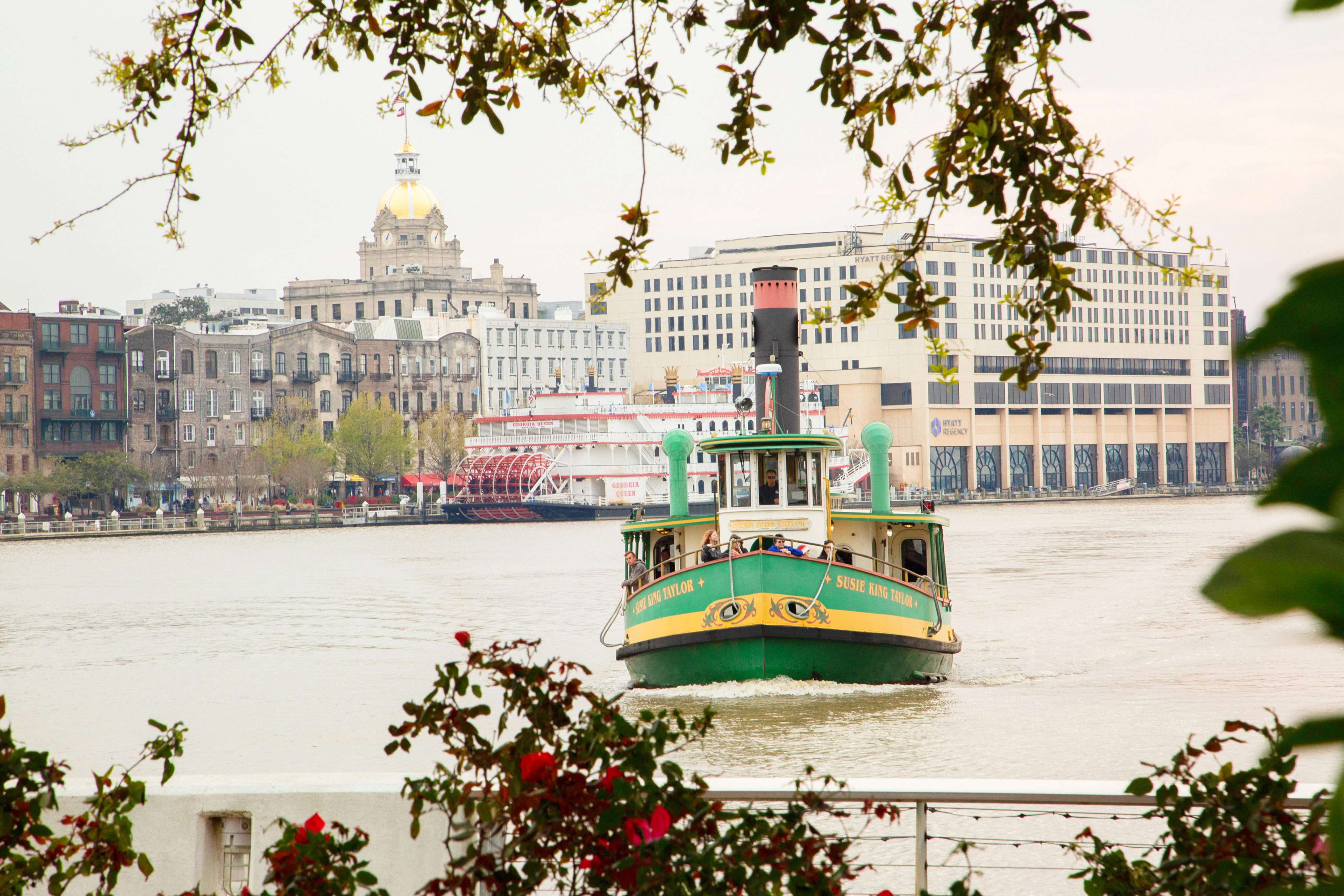 Savannah Belles Ferry, which is green with a yellow stripe, crosses the Savannah River, headed toward the camera on the Convention Center side of the river. In the background, the River Street skyline with its many buildings can be seen.